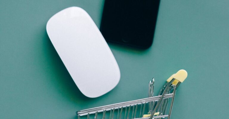 Flat lay of a miniature shopping cart, smartphone, and computer mouse on teal background.